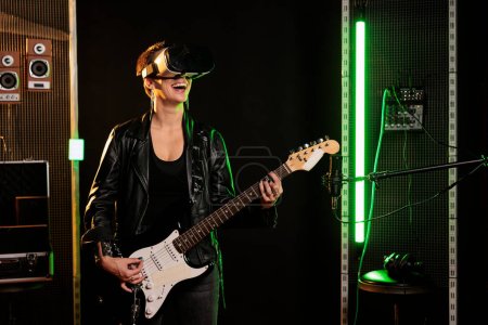Téléchargez les photos : Guitarist with virtual reality headset enjoying rock concert simulation while playing heavy metal song at electric guitar in music studio. Woman musician performing grunge album using - en image libre de droit