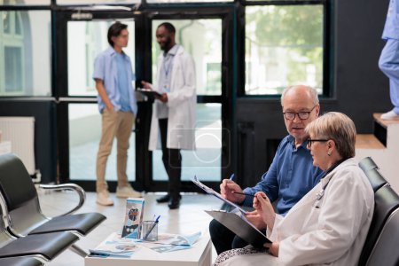 Photo for Senior doctor sitting on chair in hospital reception explaining disease diagnosis to elderly patient discussing health care treatment. People waiting to start examination during checkup visit - Royalty Free Image