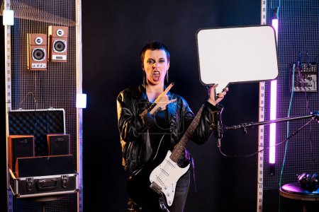 Foto de Musician showing blank placard with copy space for advertisement in music studio, person playing guitar preparing for rock concert. Rebel woman with leather outfit performing heavy metal song - Imagen libre de derechos