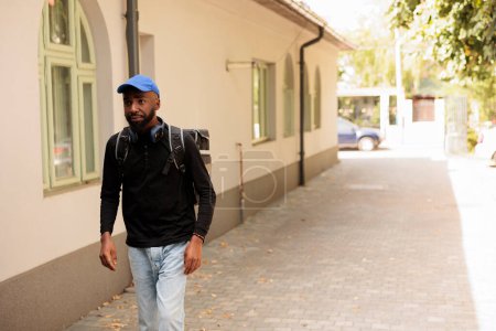 Téléchargez les photos : African american food courier walking in street, standing outdoors, front view. Deliveryman going with customer order in thermal backpack, man delivering eatery takeaway meal - en image libre de droit