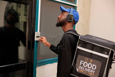 Téléchargez les photos : Office meal delivery service courier ringing building doorbell, waiting for client outdoors, side view. African american deliveryman with backpack delivering takeaway restaurant lunch - en image libre de droit