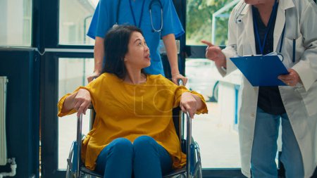 Téléchargez les photos : Femme asiatique handicapée physique recevant de l'aide d'une infirmière, ayant rendez-vous de visite de contrôle avec un médecin. Assistant aidant le patient aux prises avec une déficience chronique. - en image libre de droit