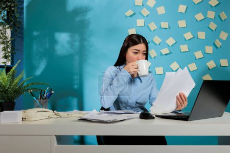 Foto de Corporate female auditor sitting at office desk analyzing financial reports of startup company. Professional woman drinking cup of coffee while comparing digital information and printed paperwork. - Imagen libre de derechos