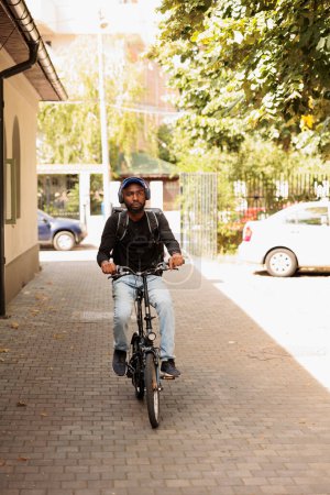 Foto de Courier in headphones riding bike on street at sunny day, looking at camera, front view. African american deliveryman with customer order in thermal bag portrait, man delivering eatery meal - Imagen libre de derechos