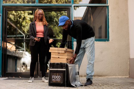Téléchargez les photos : Pizzeria delivery service, courier passing african american woman pizza boxes stack, man taking out takeaway order from thermal backpack. Deliveryman giving client fastfood - en image libre de droit