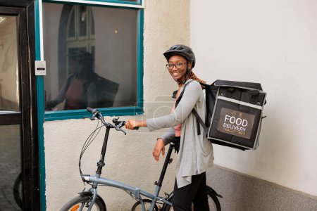 Photo for Smiling woman delivering food, courier looking at camera, waiting for customer near office building entrance outdoors. Restaurant takeaway meal delivery service, girl standing in front of door - Royalty Free Image