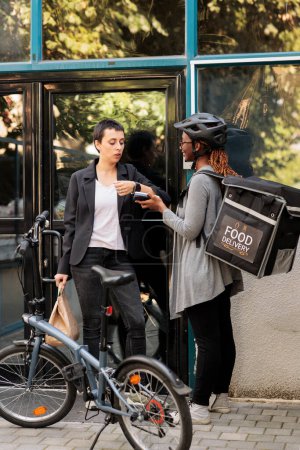 Foto de Office employee paying for delivery with smart watch contactless payment transaction. African american courier holding pos terminal, standing near company building outdoors - Imagen libre de derechos