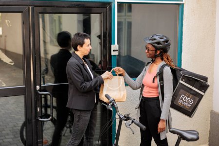Téléchargez les photos : Food delivery service, young smiling courier giving client package. African american woman holding restaurant takeaway meal order, customer receiving lunch paper bag in front of company building - en image libre de droit