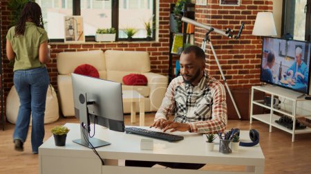 Téléchargez les photos : Afro-Américain travaillant sur ordinateur au bureau, faisant des tâches à distance à la maison avec sa petite amie. Jeune adulte utilisant l'application Web en ligne pour planifier les données financières pour le rapport de l'entreprise, envoyer un courriel. - en image libre de droit