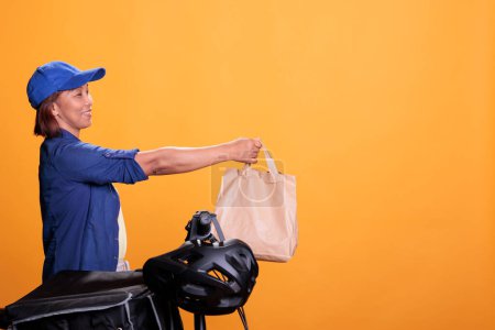 Foto de Senior takeaway delivery employee delivering takeout food in recycled paper bags to client, using bike as transportation. Restaurant worker wearing blue t shirt and cap while working as courier - Imagen libre de derechos