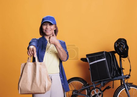 Photo for Smiling deliverywoman doing thumbs up gesture while delivering take away food to customers, using paper bags. Restaurant worker bringing orders with bike. Takeout food service and concept - Royalty Free Image