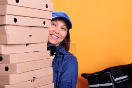 Téléchargez les photos : Portrait of asian elderly delivery worker holding pizza boxes, delivering to client during lunch time. Pizzeria employee hidden behind a large stack of takeout food order. Take out food concept - en image libre de droit