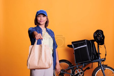 Foto de Pizzeria delivery worker wearing blue uniform while delivering fast food order to customer during lunch time. Restaurant employee standing in studio with yellow background. Takeout service concept - Imagen libre de derechos
