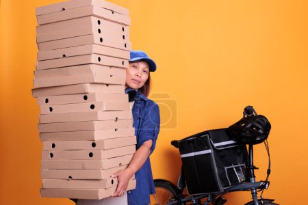 Téléchargez les photos : Large stack of pizza being carried by delivery woman in uniform for one of restaurant clients during lunk time. Elderly asian employee deliverying food order with bike, take away concept - en image libre de droit