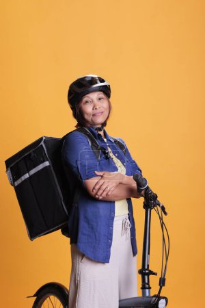 Téléchargez les photos : Smiling takeaway delivery person carrying thermal takeout food backpack, delivering pizza meal from restaurant. Asian bicyclist wearing helmet during deliver job, home delivery service - en image libre de droit