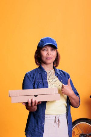 Téléchargez les photos : Asian pizzeria delivery employee doing thumbs up gesture while delivering carton flatbox with pizza to customers. Restaurant worker bringing orders with bike. Take out food service and concept - en image libre de droit