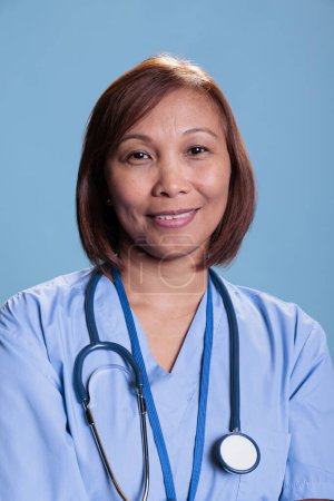 Téléchargez les photos : Smiling practitioner nurse wearing stethoscope and blue coat during medical appointment working in health care industry. Asian assistant smiling at camera standing in studio with blue background - en image libre de droit