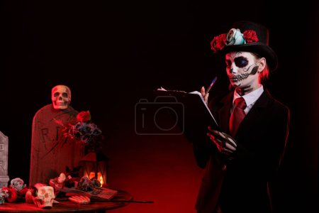 Photo for Santa muerte taking notes on clipboard files, wearing halloween dios de los muertos costume and make up. Woman celebrating la cavalera catrina on day of the dead mexican holiday in studio. - Royalty Free Image