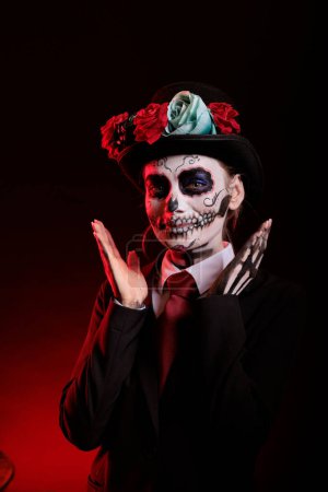 Foto de Smiling woman in costume with skull make up celebrating traditional mexican holiday with goddess of death costume. La cavalera catrina culture celebration with skull body art over black background. - Imagen libre de derechos