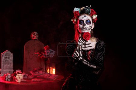 Foto de Glamour goddess of death posing with rose in studio, wearing flowers crown and santa muerte halloween costume. Looking like la cavalera catrina with body art and carnival look. - Imagen libre de derechos