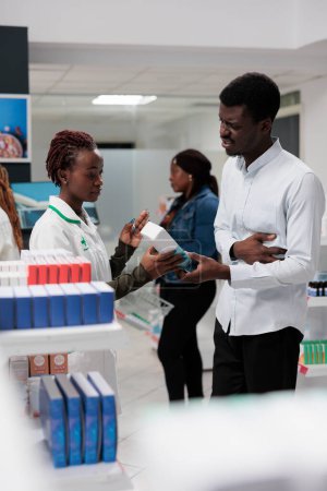 Téléchargez les photos : Client buying stomach pain medication, pharmacy consultant and buyer talking. African american customer with stomachache standing in drugstore, chatting with pharmacist, all black team - en image libre de droit