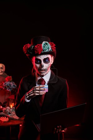 Photo for Woman in lady of death costume shopping online with credit card and laptop, wearing suit and hat with skull make up. Looking like goddess of death and santa muerte, dios de los muertos. - Royalty Free Image