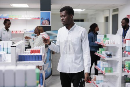 Téléchargez les photos : African american man buying heart supplements in drugstore, reading instruction on tablets package, standing in pharmacy aisle. Client choosing vitamins, pharmacists in background, all black team - en image libre de droit