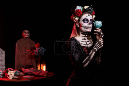 Foto de Creepy santa muerte posing with roses over black background, looking like goddess of death. Beautiful woman with skull make up and body art, festival holiday celebration in studio. - Imagen libre de derechos