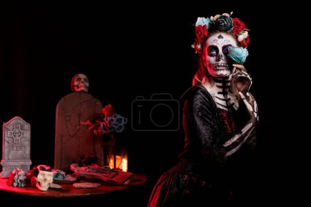 Foto de Beautiful lady dressed as catrina skull in studio, wearing traditional santa muerte holy costume with black and white make up or body art. Holding roses and having flowers crown. - Imagen libre de derechos