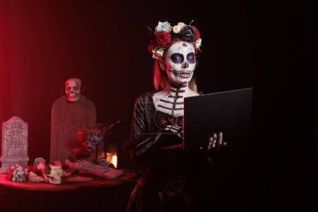 Photo for Creepy lady working on laptop in studio, checking internet website online on wireless pc. Santa muerte model wearing halloween costume and dios de los muertos skull make up on celebration. - Royalty Free Image