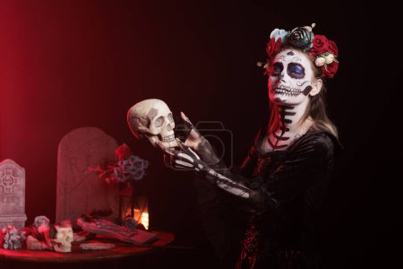 Photo for Lady of death holding smartphone for skull, acting creepy like santa muerte on mexican holiday ritual. Wearing black and white make up to celebrate dios de los muertos halloween tradition. - Royalty Free Image