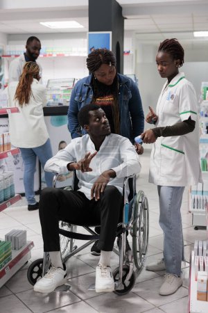 Téléchargez les photos : Pharmacist explaining african american customer in wheelchair vitamins instruction in drugstore. Man with physical impairment and social worker buying nutritional supplements, all black team - en image libre de droit