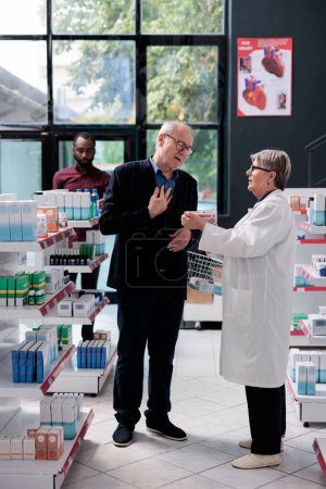 Foto de Senior specialist explaining cardiology pills treatment to old man customer, holding box of medicine and read medical leaflet. Client asking about pharmaceutics products in pharmacy shop. - Imagen libre de derechos