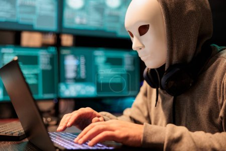 Foto de Dangerous impostor with masked identity hacking server, acting mysterious and trying to break into computer system. Hacker wearing mask and hood to steal online database information. - Imagen libre de derechos