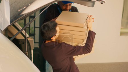 Photo for Food delivery worker giving pizza boxes to clients at office entrance, delivering big pizzeria order from car trunk. Taking lunch meal packages out of automobile to deliver takeout. Handheld shot. - Royalty Free Image