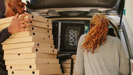 Foto de Una mensajera afroamericana sacando un montón de cajas de pizza del maletero del vehículo, entregando una gran orden de comida de pizzería a los clientes en la puerta principal. Uso de coche para entregar pila de fastfood. - Imagen libre de derechos
