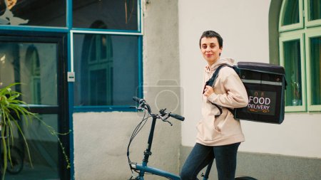 Photo for Woman working as takeaway carrier with bicycle, riding bike and carrying backpack with food package. Delivery service employee delivering fastfood restaurant meal order, sunny day. - Royalty Free Image