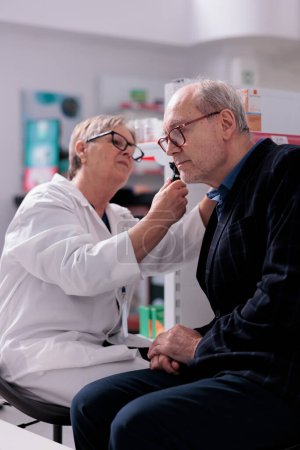 Photo for Pharmacy employee checking old man ear with otoscope and examining for infection. Elderly patient getting otoscopy consultation from pharmaceutical specialist in drugstore - Royalty Free Image