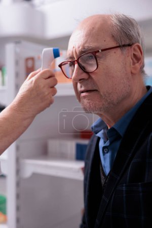 Foto de Elderly man getting temperature measuring in pharmacy store. Medical worker examining old patient with non contact infrared thermometer and doing health check up in apothecary - Imagen libre de derechos