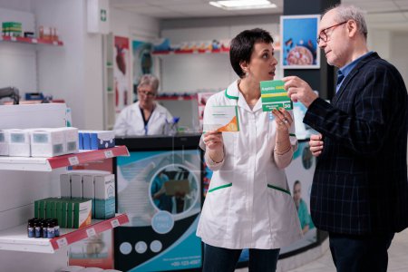 Téléchargez les photos : Elderly man asking pharmacist for help to choose pills and discussing vitamin c benefits. Young woman pharmacy consultant holding aspirin package and explaining asa dosage instruction - en image libre de droit