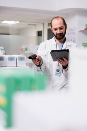Foto de Pharmacist checking medication list on tablet computer while scanning pills packages bar code using store scanner in pharmacy. Drugstore employee is responsible for organizing and labeling the drugs - Imagen libre de derechos