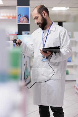 Foto de Pharmacist in white coat holding tablet computer typing vitamins barcode while doing drugs inventory in pharmacy. Drugstore employee is trained to recognize the various types of pills and packages - Imagen libre de derechos