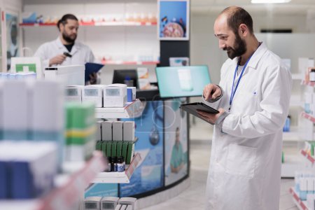 Foto de Drugstore worker looking at pills packages typing medicaments name on tablet computer during inventory in pharmacy. Pharmacist is an expert in pharmaceuticals and is able to provide guidance - Imagen libre de derechos