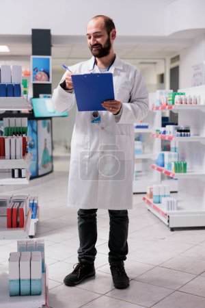 Photo for Drugstore worker holding clipboard while checking vitamins packages writing medication name on papers, working in pharmacy. Pharmacist is responsible for maintaining the inventory of pills - Royalty Free Image