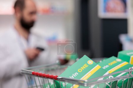 Photo for Selective focus on basket full with supplements packages standing on counter desk in pharmacy. In background pharmacist scanning products, being responsible for maintaining the inventory of pills - Royalty Free Image
