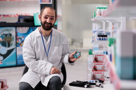 Foto de Pharmacist standing on chair in drugstore holding medical glucometer looking at camera while waiting for customers. Pharmacy equipped with pharmaceutical products, vitamins and pills - Imagen libre de derechos
