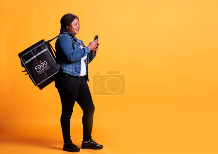 Téléchargez les photos : Full body of pizzeria courier carrying takeaway thermal backpack while checking client adreess on smartphone before start deliver food order during lunch time. Takeout service concept - en image libre de droit