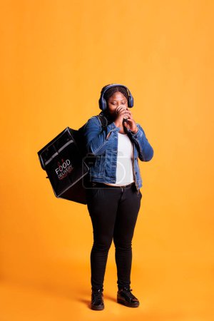 Photo for Deliverywoman with headphone drinking coffee while delivering takeaway food order to clients during lunch time in studio. African american restaurant courier carrying takeout thermal backpack - Royalty Free Image