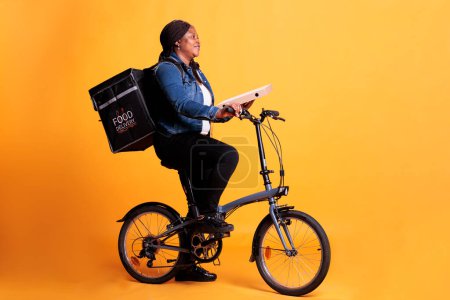 Photo for Full body of woman courier holding pizza cardboard while riding bike delivering takeaway meal to client during lunch time. Restaurant worker standing in studio with yellow background - Royalty Free Image