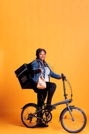 Téléchargez les photos : African american pizzeria courier holding brown paper bag delivering takeaway food meal to client during lunch time standing in studio with yellow background. Takeout service concept - en image libre de droit